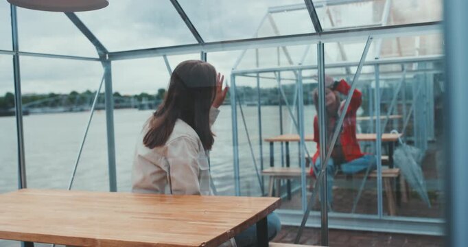 Two young women sitting in separate transparent cabins with hands on glass, looking at each other
