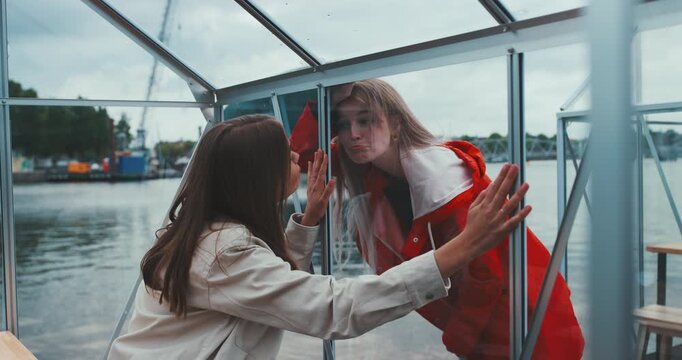 Beautiful young woman outside glass cabin looking in with sad calm expression, friend inside with hands on glass
