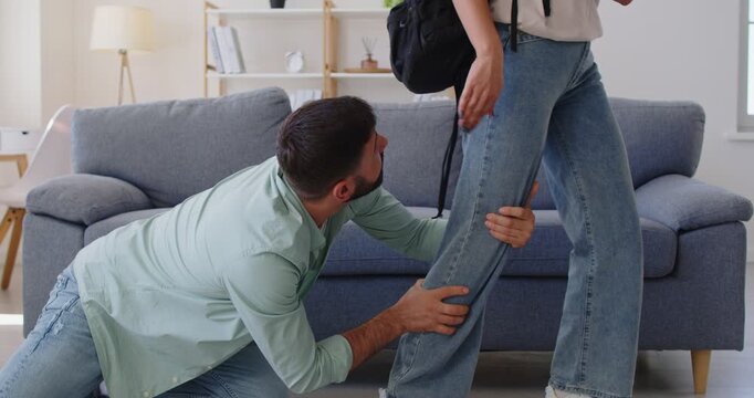Man kneeling begging to forgive, pleading young husband and wife with backpack leaving home, conflict and breakup in couple. Clingy male partner holding leg of girl with grip and drama in living room