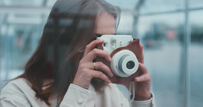 Young woman inside glass cabin taking photo with instant camera, smiling, view from outside
