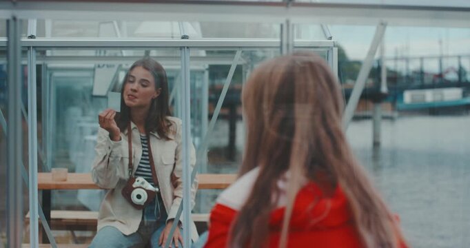 Young woman inside glass cabin looking at instant photo while friend stands outside seen from behind
