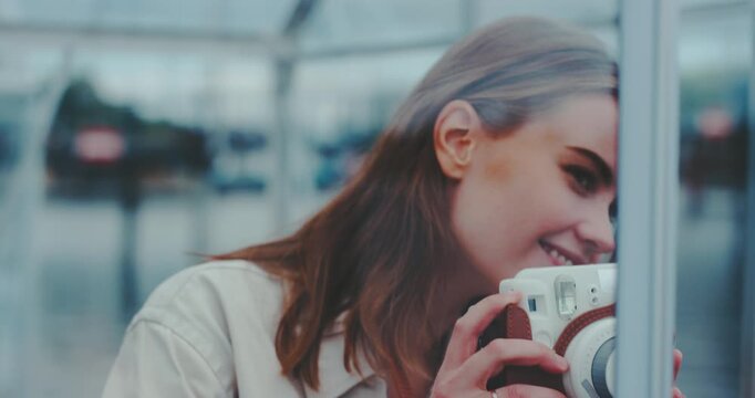 Beautiful young woman inside glass cabin aiming instant camera and framing shot, view through glass
