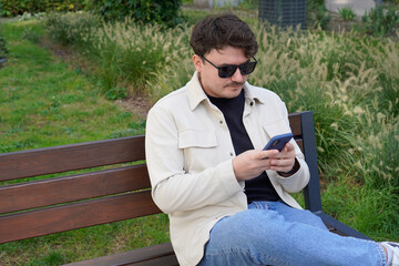 Fototapeta premium A Caucasian man with glasses sits on a bench and types a message on his phone
