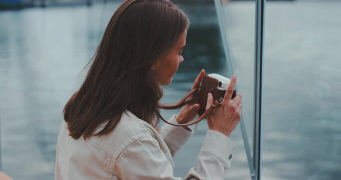 Young woman inside glass cabin seen from behind holding instant camera and framing shot
