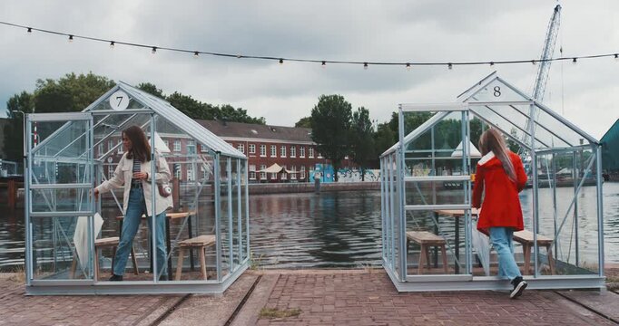 Two young women entering separate glass cabins and sitting down
