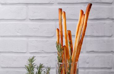 Grissini made of wheat flour with parmesan, rosemary and olive oil served in a glass. Breadsticks, Italian crispy snack, against the grey brick wall. Copy space