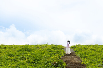 Fototapeta premium Asian woman standing on outdoor stairway with green grass and cloudy sky background. Minimal lifestyle travel concept with freedom and nature