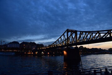 The Eiserner Steg, an iron bridge in Frankfurt, Germany, illuminated at dusk over the Main River with the city skyline in the background.