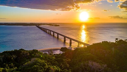 Obraz premium Aerial view of a long bridge spanning across ocean water, with the setting sun casting a golden glow, and an island