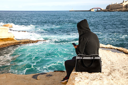 On the rugged Maltese shoreline, a hooded figure holds a smartphone, trying to make a call, with a black bag nearby, capturing a quiet moment against the coastal backdrop.