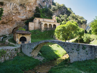 Fototapeta premium Mountain Monastery with Goat on Bridge - A black goat stands on a stone bridge over a small river, with a historic monastery built into the side of a steep, rocky cliff in the background.