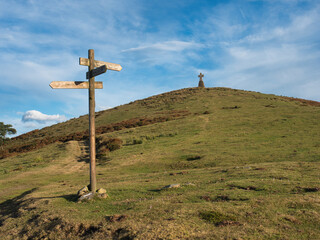 Trail Signs on a Grassy Hill - A wooden trail signpost stands on a grassy, sunlit hill with a rocky cross at the summit, guiding hikers under a wide, blue sky with wispy clouds. © jmag.foto