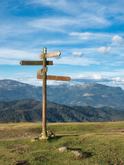 Trail Signs on a Grassy Hill - A wooden trail signpost stands on a grassy, sunlit hill with a rocky cross at the summit, guiding hikers under a wide, blue sky with wispy clouds. © jmag.foto