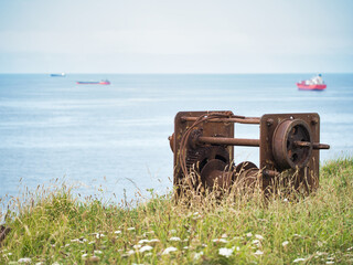 Fototapeta premium A close-up, eye-level shot of a large, weathered, and rusted metal winch on a grassy clifftop, with the calm blue sea and cargo ships visible in the distance.