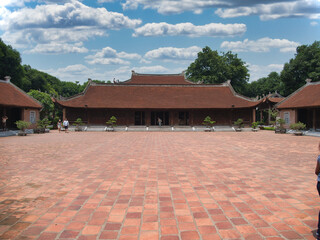 Fototapeta premium Vietnamese temple with large brick courtyard - The front view of a traditional Vietnamese temple with a large, empty brick courtyard, surrounded by buildings with curved roofs and lush trees.