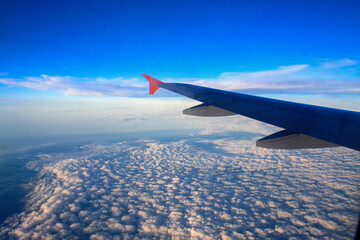 Obraz premium A stunning aerial view from an airplane window shows a wing gliding above a vast blanket of clouds, with a bright blue sky and soft white formations stretching endlessly on the horizon.