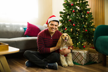 Obraz premium Portrait of a Hispanic man hugging his dog wearing christmas clothes by the tree