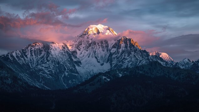 Majestic snow-capped mountain peaks are illuminated by the warm glow of the rising or setting sun, as pastel-colored clouds gently drift above the rugged terrain, casting a soft, ethereal light over.