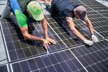 Workers building photovoltaic solar panel system on rooftop of house. Men engineers installing solar module with help of wrench outdoors. Alternative and renewable energy generation concept.
