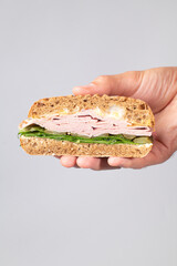 Sandwich with ham, with pickles and lettuce leaves in hand on grey background.