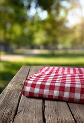 Red and white checkered picnic cloth folded on rustic wooden table outdoors, blurred green park background