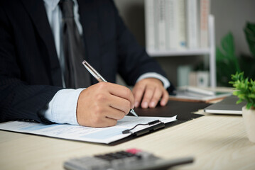 Businessman in suit signing contract document at office desk. Closeup hand holding pen for business agreement, partnership deal, legal commitment, finance investment, or insurance policy approval.