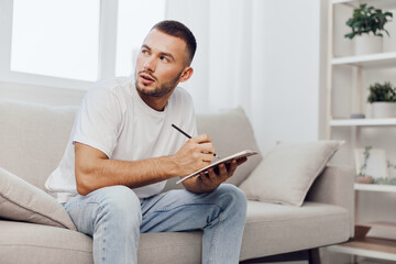 Thoughtful man sitting on a sofa, writing notes in a notebook, dressed casually in a white t-shirt and jeans, creating ideas for future projects.
