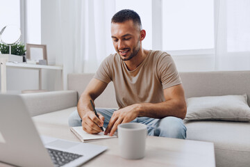 Engaged man writing in a notebook, sitting on a cozy sofa with a laptop nearby, conveying dedication and productivity in a bright, airy living space.