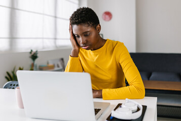 Worried young african woman using laptop at home. Unhappy black female reading bad news on internet...