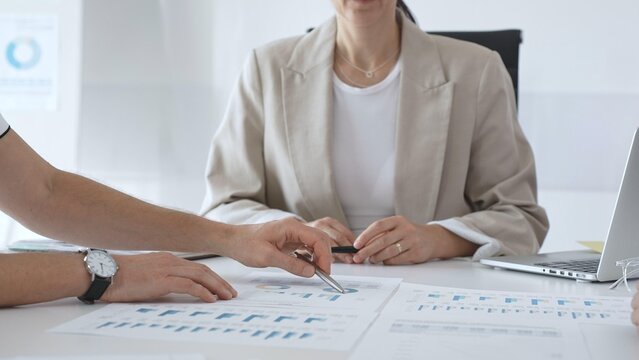 Professional businesspeople collaborating on financial analysis, female executive gesturing toward charts while presenting strategic insights to team members in corporate workspace - Powered by Adobe