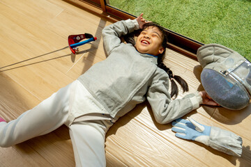 Close up of Asian girl with fencing sport uniform lie down on the floor and action like happy with smiling warm light from glass door of balcony.