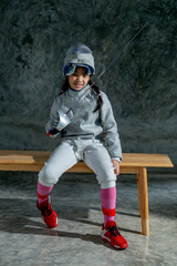 Vertical image of girl wear fencing sport uniform also hold epee and sit on wooden bench with smiling to relax after practice for the competition.