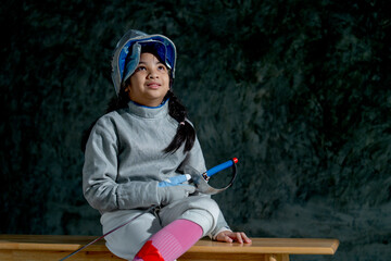 Close up Asian girl with fencing sport uniform hold epee and look up to her left side also sit on wooden bench and smiling with happiness.