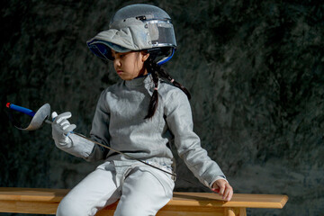 Close up Asian girl with fencing sport uniform hold epee and look at it also sit on wooden bench look like thinking about the competition.