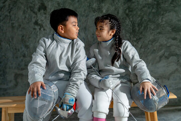 Asian brother and sister with fencing sport uniform sit beside on wooden bench and look to each other and smile also relax after practice for competition.