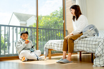 Asian mother sit on bed and look to his son who enjoy eat an apple also sit on the floor during practice fencing sport in bedroom with soft morning light and happiness.