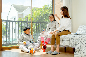 Asian mother sit on bed next to her daughter and touch hair of her daughter and boy sit on floor during practice fencing sport in bedroom with soft morning light and happiness.