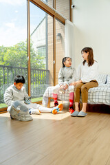 Vertical image Asian mother sit on bed next to her daughter and talk to her daughter and boy sit on floor during practice fencing sport in bedroom with soft morning light and happiness.