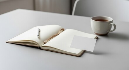 Naklejka premium Open notebook with blank pages sits on a gray table next to a cup of tea and a business card in a minimalist and calm setting.