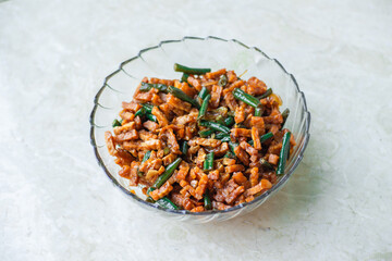 A glass bowl of Orek Tempeh, a traditional Indonesian sweet and spicy stir-fry with tempeh and green beans, on a white background