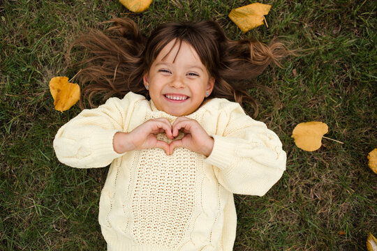 Smiling kid girl 5-6 year old lying on green grass with fallen leaves making heart shape with hands top view looking at camera. Child wearing knitted yellow sweater. Autumn season.