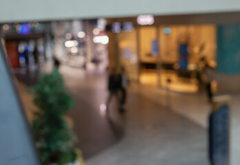 abstract, downward view of a blurred shopping mall corridor with a figure walking past a small potted tree, conveying urban life, commerce, and motion
