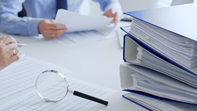 Accountants are analyzing financial reports with magnifying glass and documents, performing accounting and auditing tasks in the office, close up. Audit and taxes in business