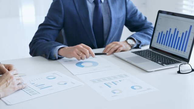 Business colleagues or partners are discussing financial data on a laptop and printed documents while analyzing charts and market trends at office table. Businesspeople and finance concept - Powered by Adobe