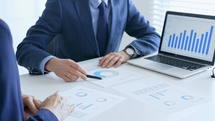 Business colleagues or partners are discussing financial data on a laptop and printed documents while analyzing charts and market trends at office table. Businesspeople and finance concept