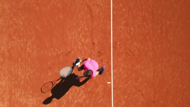 Tennis player in pink shirt practices on clay court, dynamic movement and focus captured from aerial shot, highlighting shadow and dedication to skill development.