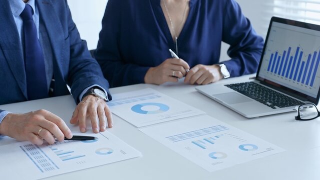 Business team analyzing financial data on a laptop and printed documents, pointing at charts and graphs during a meeting in bright office. Businesspeople and finance concept