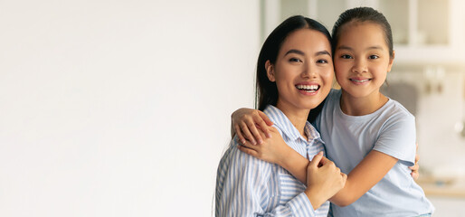 Portrait of young asian mother and cute little daughter posing in kitchen interior, pretty girl hugging her mom and smiling at camera, mommy and her child having fun at home