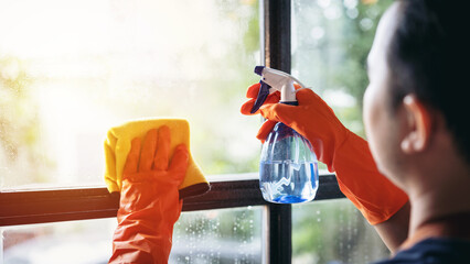 Housekeeper man in orange rubber gloves using a clean spray and a duster while cleaning on window...