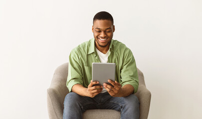 Cheerful Black Guy Using Tablet Computer Browsing Internet Or Watching Movie Online Sitting In Chair Over Gray Studio Background. Technology, Internet And Gadgets Concept